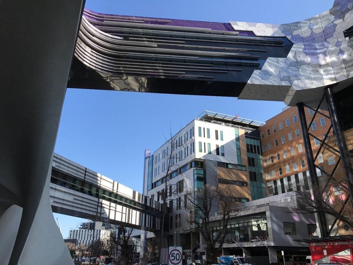 Looking up from a hospital precinct street to glazed bridges linking the buildings overhead, under a clear blue sky.